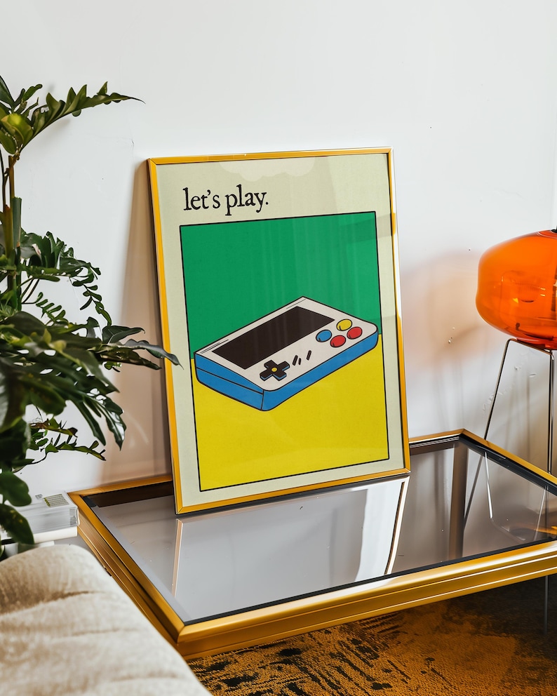 May include: A framed poster with a yellow, green, and blue color scheme. The poster features a cartoon illustration of a handheld video game console with the text "Let's play."