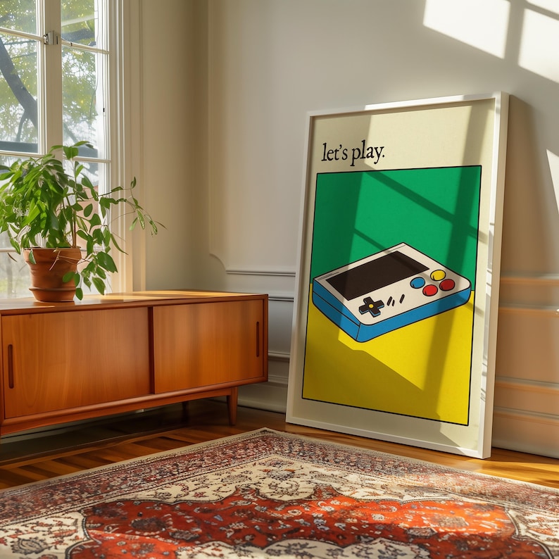 May include: A white framed poster with a green, yellow, and blue illustration of a handheld video game console. The text "Let's play." is printed in black at the top of the poster.
