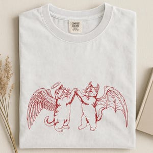May include: White t-shirt featuring a red graphic of two cats, one with angel wings and a halo, the other with devil wings and horns, holding hands. The shirt has a crew neck and a Comfort Colors tag.