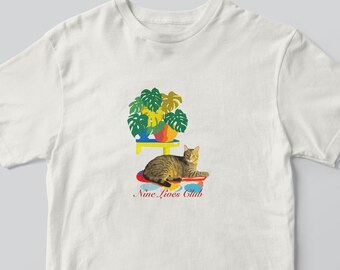 May include: White t-shirt featuring a tabby cat lying on a colorful rug. Above the cat is a yellow table with a potted plant. The text "Nine Lives Club" is below the cat.