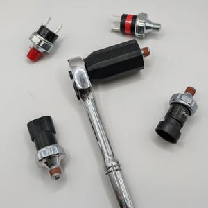 May include: An assortment of automotive pressure sensors and a chrome ratchet wrench are arranged on a white surface. The sensors have black and silver bodies with red or silver accents. One sensor is labeled "PSI".