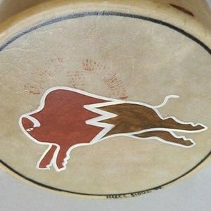 May include: A hand-painted drumhead featuring a stylized bison in shades of brown and red, outlined in white, against a tan background. The design is enclosed by a black border. The drumhead is part of a musical instrument.