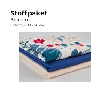 以下が含まれることがあります： 3枚の生地のスタック。一番上の生地は、クリーム色の背景に赤い花と青い葉のフローラルパターンです。真ん中の生地は青色で、一番下の生地はクリーム色です。テキストは「Stoffpaket Blumen 3 Stoffe je 30 x 50 cm」と書かれています。