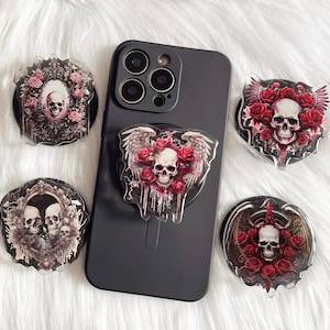 May include: Six phone grips with different skull and rose designs. The grips are made of clear plastic and have a black background. The skulls are white and the roses are red.