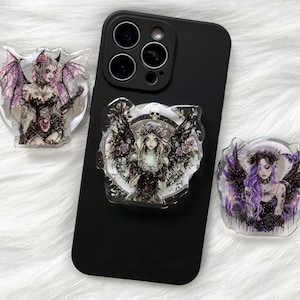 May include: Black smartphone with three decorative phone grips featuring gothic angel illustrations. The grips have wings, floral accents, and detailed artwork in shades of purple, black, and white. The phone rests on a white, fluffy surface.