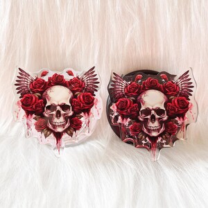 May include: Two clear acrylic phone grips with a skull and red roses design. The skull is white with black eyes and a black mouth. The roses are red with green leaves. The phone grips are shaped like a heart.