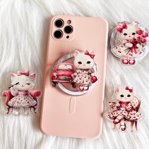 May include: A pink phone case with a pop socket featuring a cartoon cat in a dress, a red car, and other cat-themed accessories. The accessories include a cat holding a makeup brush, a cat holding a bouquet of roses, and a cat sitting in a chair.