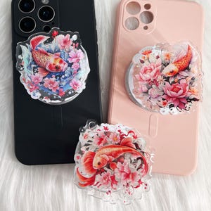 May include: Clear acrylic phone grips featuring koi fish and floral designs. The koi fish are orange and red, surrounded by pink and blue flowers. The grips are attached to black and pink phone cases.