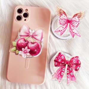 May include: Three pink and white phone grips with different designs. One features a pink bow with cherries, another has a pink bow with wings, and the last one has a pink bow with a skull and crossbones pattern.