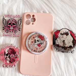 May include: Four phone accessories with a skull and floral design. The accessories are a phone case, a pop socket, a phone ring, and a phone charm. The phone case is pink and has a clear back. The pop socket is clear and has a skull and floral design. The phone ring is clear and has a skull and floral design. The phone charm is clear and has a skull and floral design. The text on the accessories says "Wild spirit Sweet Soul", "Dead inside 2 but it's Valentines", "Beautiful Crazy", and "Bite Me".