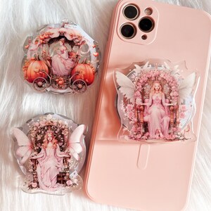 May include: Three phone grips with a fairytale theme. The first grip features a carriage with a princess inside, the second grip features a princess sitting on a throne, and the third grip features a fairy with wings.