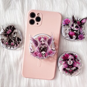 May include: Four phone grips with different designs featuring gothic and spooky characters. Each grip has a pink, white, and black color scheme with floral accents.