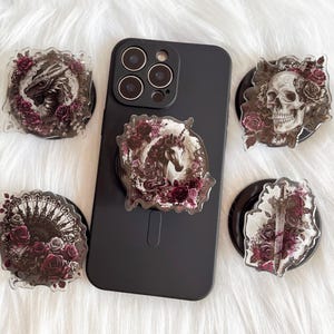 May include: Five phone grips with different designs. Each grip features a black and white illustration of a mythical creature surrounded by dark red roses. The designs include a dragon, a unicorn, a skull, a sword, and a fan.
