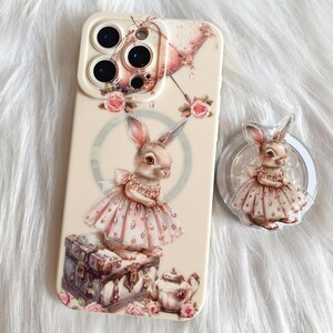 以下が含まれることがあります： 白い携帯電話ケースに、ピンクと白のウサギがドレスを着てティーカップを持っています。ケースには3つの円形のカメラレンズが付いています。背景は薄いピンクと白の花柄です。