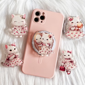 Könnte beinhalten: Ein rosa Smartphone-Hülle mit einem Pop-Socket mit einem Cartoon-Katzen-Design. Mehrere katzenförmige Acryl-Anhänger umgeben das Telefon, jede trägt ein Kleid mit Kirschmuster. Die Anhänger haben einen weißen Hintergrund und sind mit rosa Schleifen und Accessoires verziert.