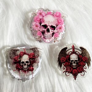 May include: Three clear acrylic charms featuring a skull with wings and flowers. The first charm has a pink skull with pink flowers. The second charm has a white skull with red flowers. The third charm has a white skull with red flowers and wings.