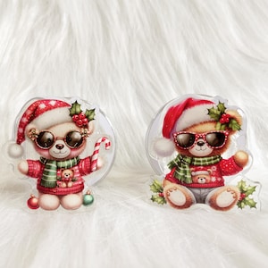 May include: Two Christmas-themed bear pacifier clips. Both bears are wearing red and white Santa hats, sunglasses, and red sweaters. One bear is holding a candy cane and the other is wearing a green scarf.