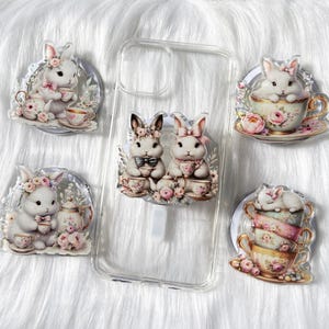May include: Four phone grips featuring white bunnies with pink bows drinking tea from teacups. The bunnies are surrounded by flowers and leaves. The phone grips are designed to be attached to the back of a phone case.
