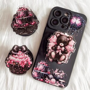 May include: Three phone grips featuring a black and pink cupcake, a black and pink cake, and a black teddy bear with pink flowers. The grips are on a black phone case with a camera lens and the text "Pro Camera".