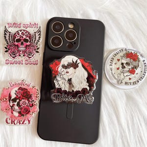May include: A black phone case with four decorative acrylic phone grips. The grips feature skull designs with roses, wings, and the phrases "Wild Spirit Sweet Soul", "Beautiful Crazy", "Bite Me", and "Dead Inside But It's Valentine's".