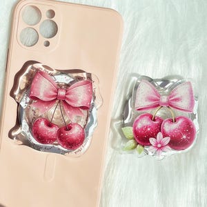 May include: A pink phone case with a clear, decorative phone grip featuring a pink bow and two red cherries. A matching cherry and bow phone grip is also shown. The phone case is light pink.