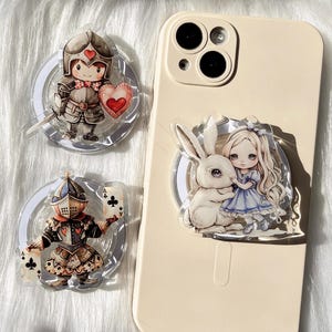 May include: Clear acrylic phone grips featuring cartoon illustrations. One grip depicts a knight with a heart shield and sword. Another shows a knight holding playing cards. A third grip shows Alice and the White Rabbit.