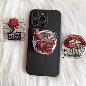 Puede incluir: Tres pegatinas para funda de teléfono con un esquema de color rojo, negro y blanco. Una pegatina dice "Guilty of Love" con una calavera y huesos cruzados. Otra pegatina dice "Hello Gorgeous" con una mano de esqueleto y labios. La tercera pegatina dice "Love" con una mano de esqueleto y un corazón a cuadros.