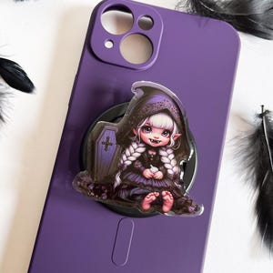 May include: A purple phone case with a clear, round phone grip featuring a cartoon illustration of a young girl with white hair, wearing a black and purple dress and a black hooded cloak. The girl is holding a small white object in her hands. The background of the illustration is a dark purple coffin with a white cross.