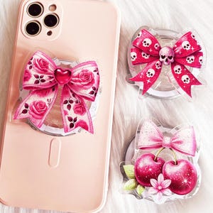May include: Three pink and white phone grips with different designs. One grip features a pink bow with a heart in the center, another grip features a pink bow with cherries and a flower, and the last grip features a pink bow with skulls.