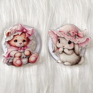 May include: Two adorable character charms, a teddy bear wearing a pink dress and a bunny wearing a pink hat. Both charms are on a clear background.