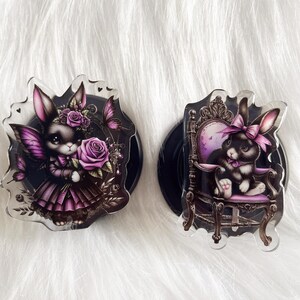 May include: Two black and purple phone grips with a rabbit design. One grip features a rabbit holding a purple rose and the other features a rabbit sitting on a throne.