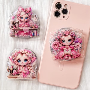 May include: Three phone grips featuring illustrations of girls with pink and white dresses and floral crowns. The girls are holding sewing tools and are surrounded by sewing-themed imagery.