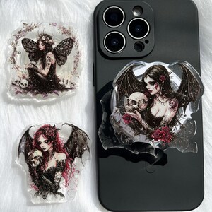 Op de afbeelding: Drie acryl telefoonhouders met gotische kunst. Eén toont een vrouw met vleermuisvleugels en een schedel, een ander een vrouw met vleugels en de derde een vrouw met rood haar en vleermuisvleugels, die een schedel vasthoudt. Op een zwarte telefoon.