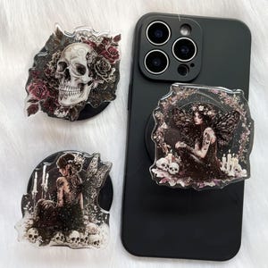 Op de afbeelding: Drie decoratieve telefoonhouders met gotische ontwerpen. Eén heeft een schedel met rozen en twee beelden een fee af met schedels en kaarsen. De houders zijn zwart met duidelijke, gedetailleerde afbeeldingen.