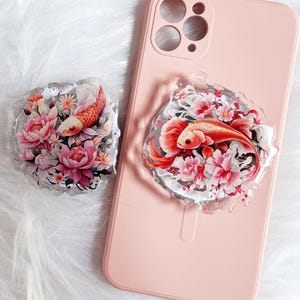 May include: A clear phone grip and a matching phone grip attached to a pink phone case. Both grips feature a koi fish design with pink flowers and foliage. The koi fish is orange and white.