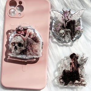 May include: A pink phone case with a clear, decorative sticker featuring a woman with a skull. Two additional stickers with similar gothic fairy designs are also visible. The stickers have a transparent background and are adorned with skulls and floral elements.