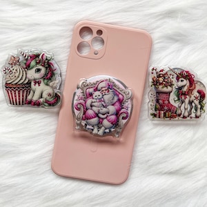 May include: A pink phone case with three decorative phone grips. The grips feature a unicorn with a cupcake, Santa Claus holding a cat, and a unicorn with a Christmas stocking. The grips have a clear, glossy finish.