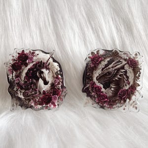 May include: Two decorative glass ornaments featuring a unicorn and a dragon. Both ornaments are surrounded by dark red roses and have a vintage aesthetic.