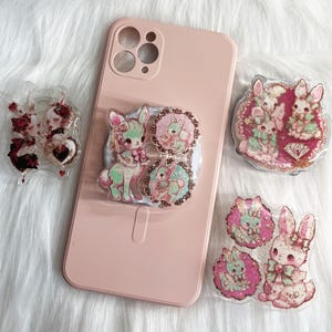 Może przedstawiać: Różowe etui na telefon z uchwytem popsocket z motywem jelenia i królika. Kilka dekoracyjnych elementów z żywicy z motywami królików i jeleni jest rozrzuconych wokół etui na telefon, prezentując pastelową paletę kolorów.