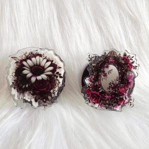 Puede incluir: Dos agarres para teléfono con diseños florales. Un agarre presenta una flor blanca con acentos rojos y el otro presenta un espejo con un diseño floral.