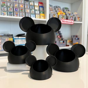 May include: Four black ceramic containers shaped like Mickey Mouse heads, with varying sizes. The containers have a smooth, matte finish and are ideal for holding small items.