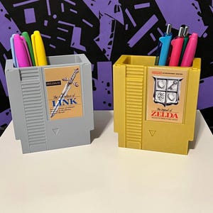 May include: Two pen holders shaped like classic video game cartridges. One is gray with "The Adventure of Link" label, the other is yellow with "The Legend of Zelda" label. Each holds colorful pens.