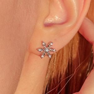 May include: A silver snowflake stud earring with small, clear crystals. The earring is worn on an ear, with the person's hand gently touching the ear. The earring is a delicate piece of jewelry.
