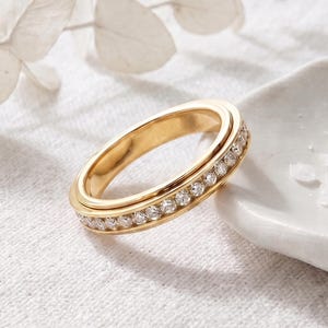 May include: A gold-toned metal ring featuring a central channel set with a continuous row of small, clear, round-cut stones. The band has a smooth, polished finish and a rounded profile, showcasing a consistent design around the entire circumference.