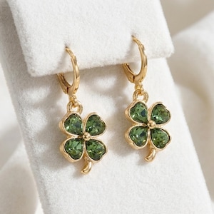 Peut inclure: Une paire de boucles d'oreilles cr&eacute;oles dor&eacute;es avec des pendentifs en forme de tr&egrave;fle &agrave; quatre feuilles. Chaque tr&egrave;fle est compos&eacute; de quatre pierres en cristal vert fonc&eacute; facett&eacute;es, serties dans une monture en m&eacute;tal dor&eacute; poli.