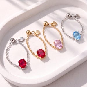 May include: Four chain rings featuring oval-cut gemstones in red, pink, and blue. Two rings are silver-toned with red and blue stones, and two are gold-toned with red and pink stones. Each ring includes a small heart charm and a spherical bead.