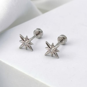 May include: A pair of silver-colored stud earrings. Each earring features a starburst design with a small, clear gemstone at the center. The earrings are displayed on a white surface.