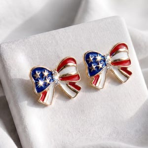 May include: A pair of bow-shaped earrings with a patriotic design. The earrings feature the colors of the American flag: red, white, and blue. The blue section has white stars, and a small crystal is in the center of each bow. The earrings have a gold-colored border.
