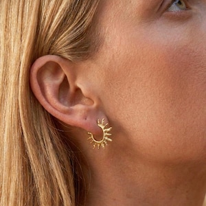 Op de afbeelding: Een gouden oorring met een gestructureerd zonnestraalontwerp met stralen die zich uitstrekken vanaf de buitenrand van de cirkelvormige band. Het metaal heeft een matte, licht onregelmatige afwerking, wat zorgt voor een opvallende, organische uitstraling.