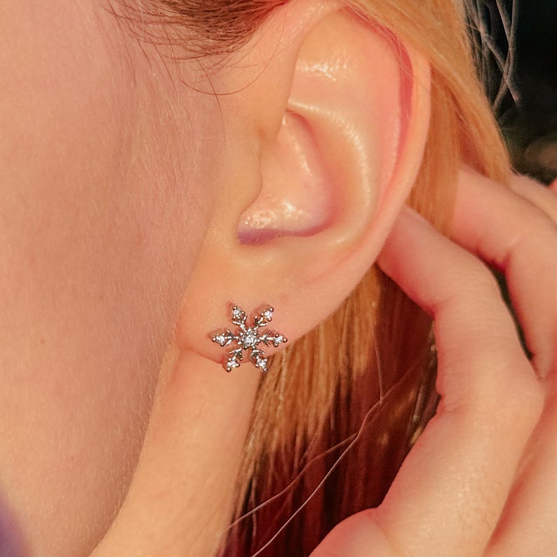 May include: A silver snowflake stud earring with small, clear crystals. The earring is worn on an ear, with the person's hand gently touching the ear. The earring is a delicate piece of jewelry.